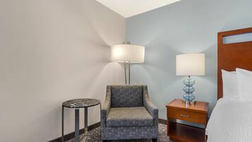 A hotel room with a grey armchair, a small round table, a bedside table with a lamp, and a bed with white bedding.