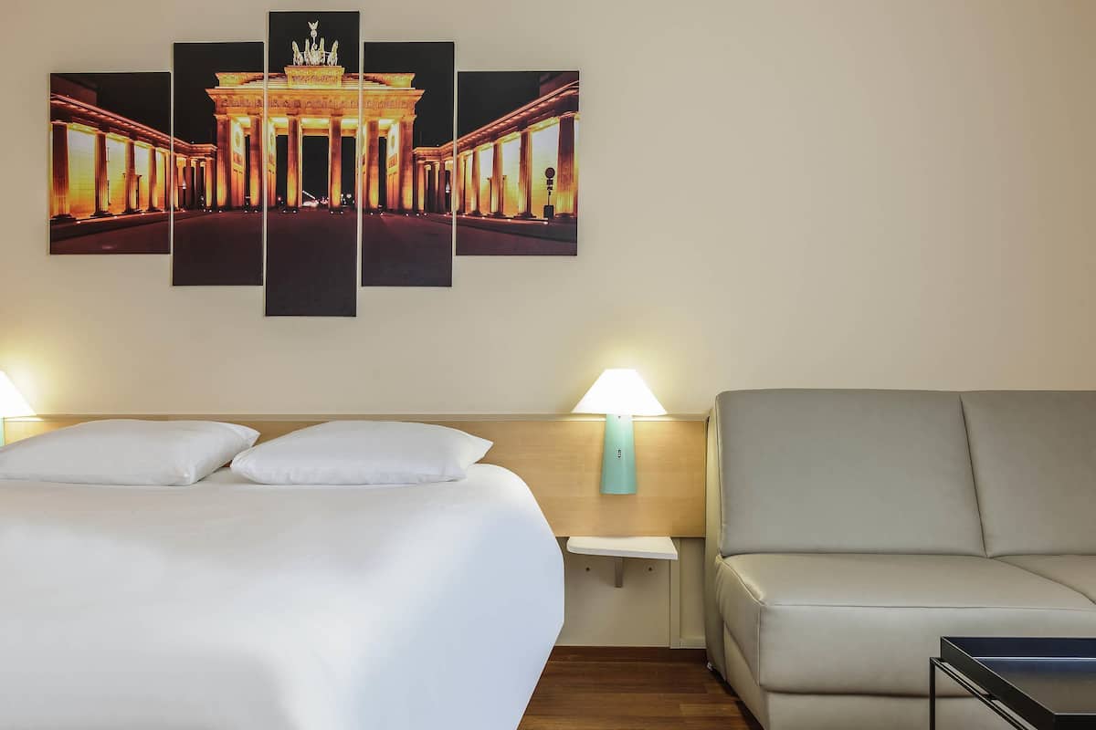 Una camera d'albergo con un letto, un divano, un comodino, una lampada e un quadro appeso al muro raffigurante un edificio.