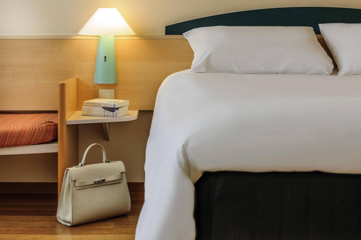 A hotel room with a neatly made bed, a bedside table with a lamp, a small shelf with books, and a handbag on the floor.