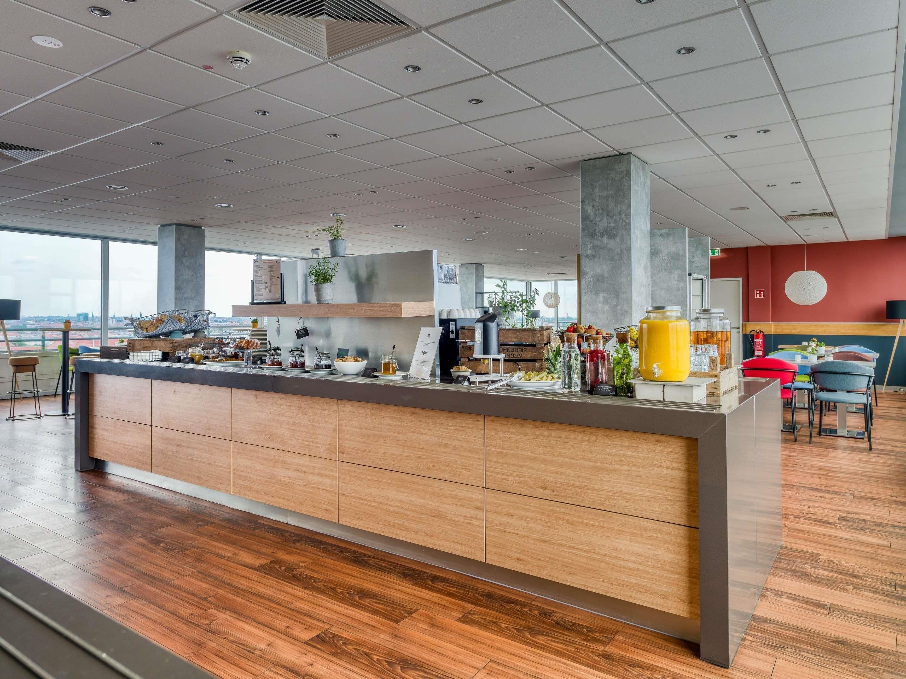 A modern breakfast bar with a wooden finish, a variety of food and drink options, and a view of the cityscape through large windows.