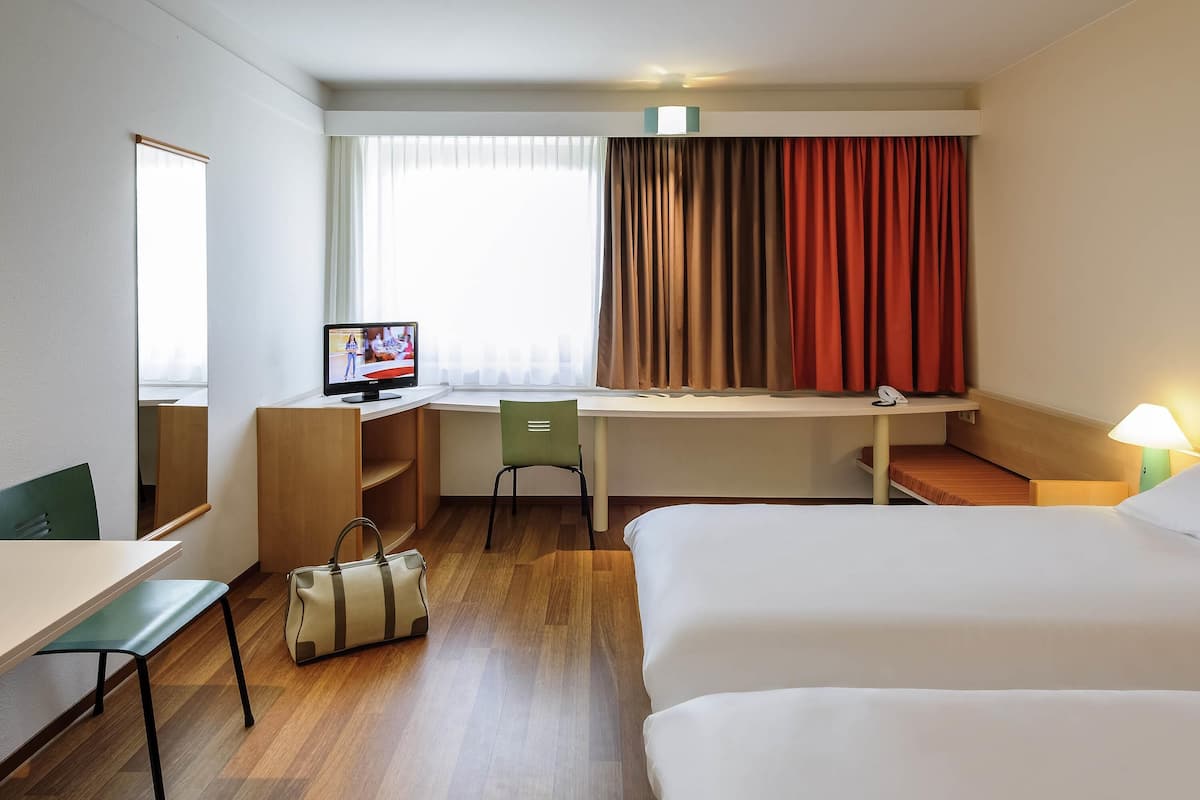 Una camera d'hotel con una scrivania, una sedia, un letto, una TV, una lampada e una borsa.