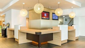 A reception area with a modern design, featuring a reception desk, multiple pendant lights, and digital screens displaying welcome messages. - ibis Berlin Messe (Berlin)