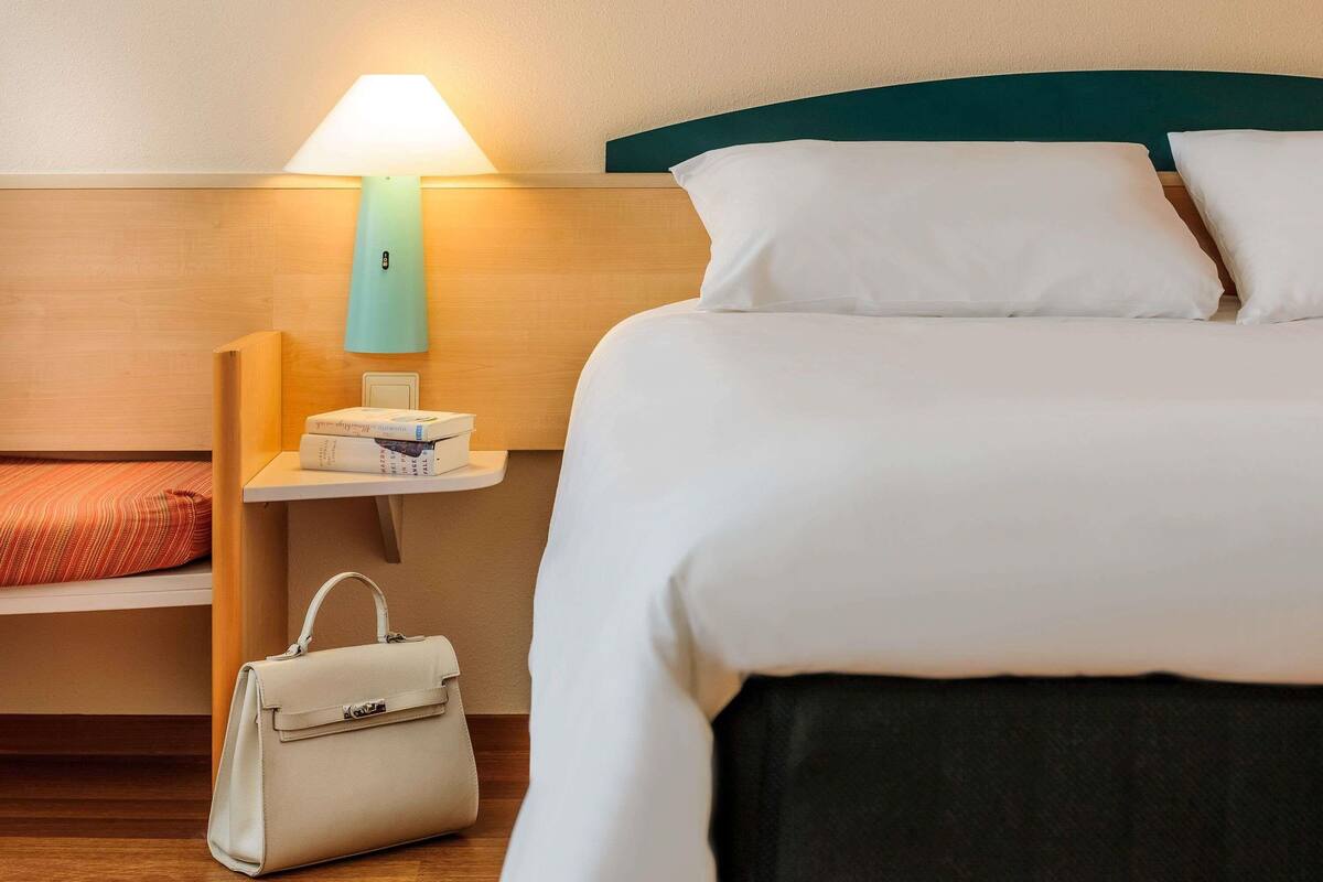 Una habitación de hotel impecable con cama, mesita de noche con lámpara, pequeña estantería con libros y un bolso de mano.