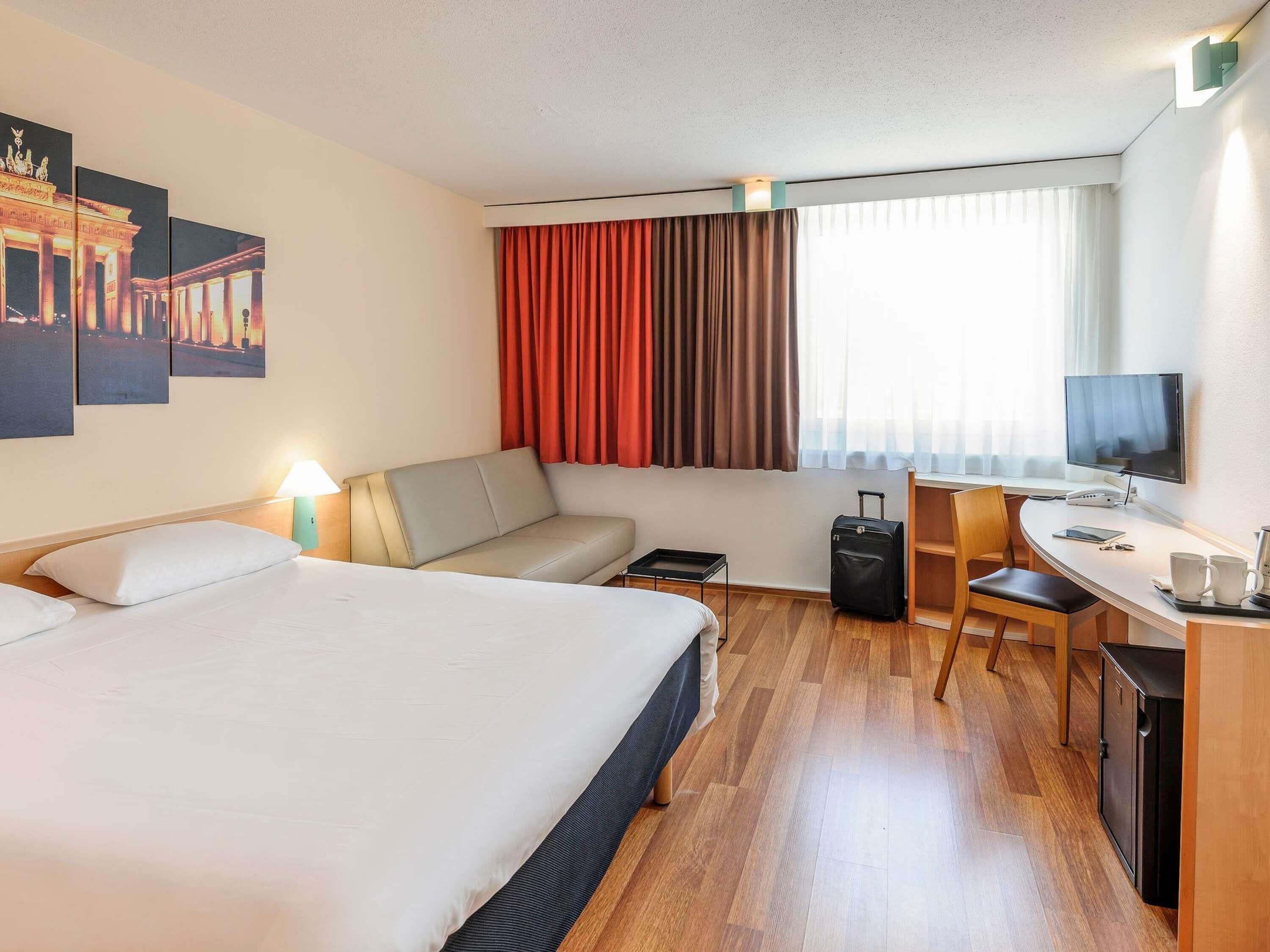 A hotel room with a bed, a sofa, a desk with a computer, a chair, and a suitcase.