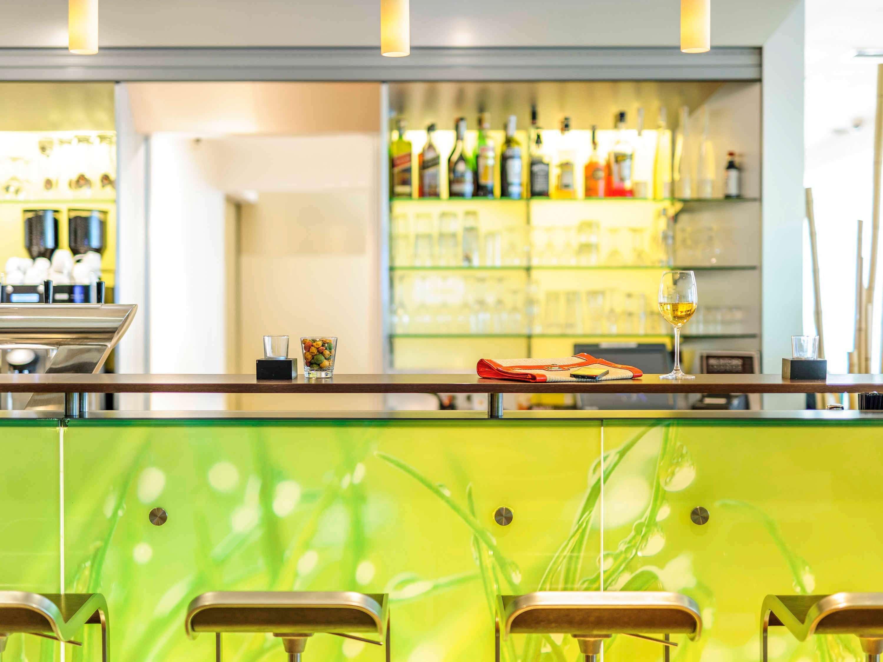 A bar area with a green wall, bar stools, and a variety of bottles and glasses on shelves.