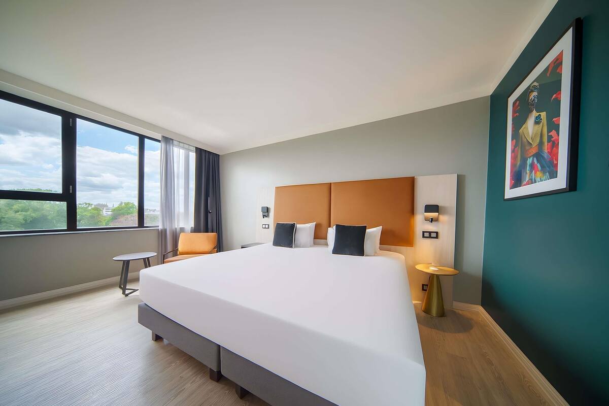 A modern hotel room with a large bed, a small table, a chair, and a view of the outdoors through large windows.