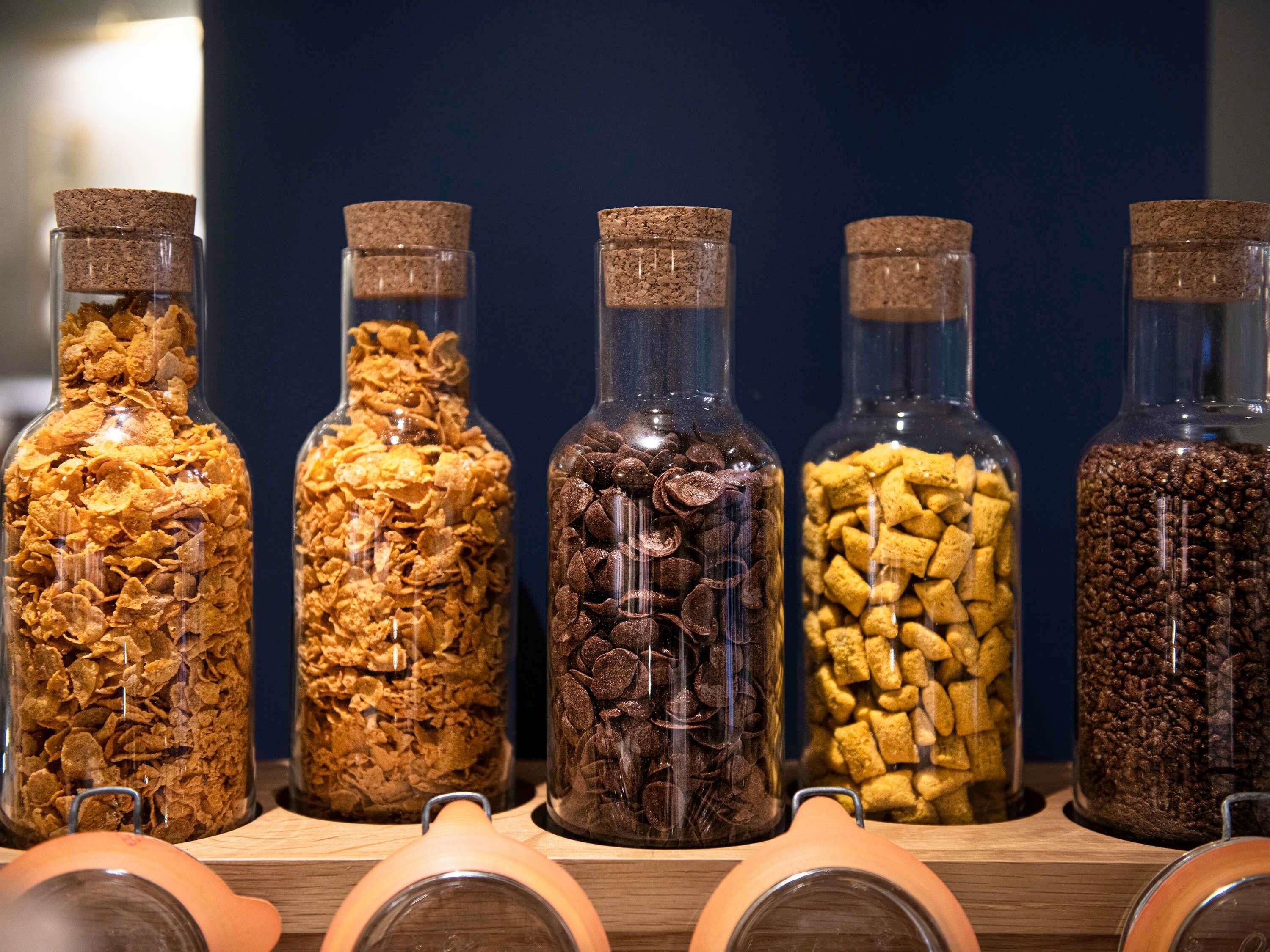 Five glass jars with cork lids containing different types of cereal.