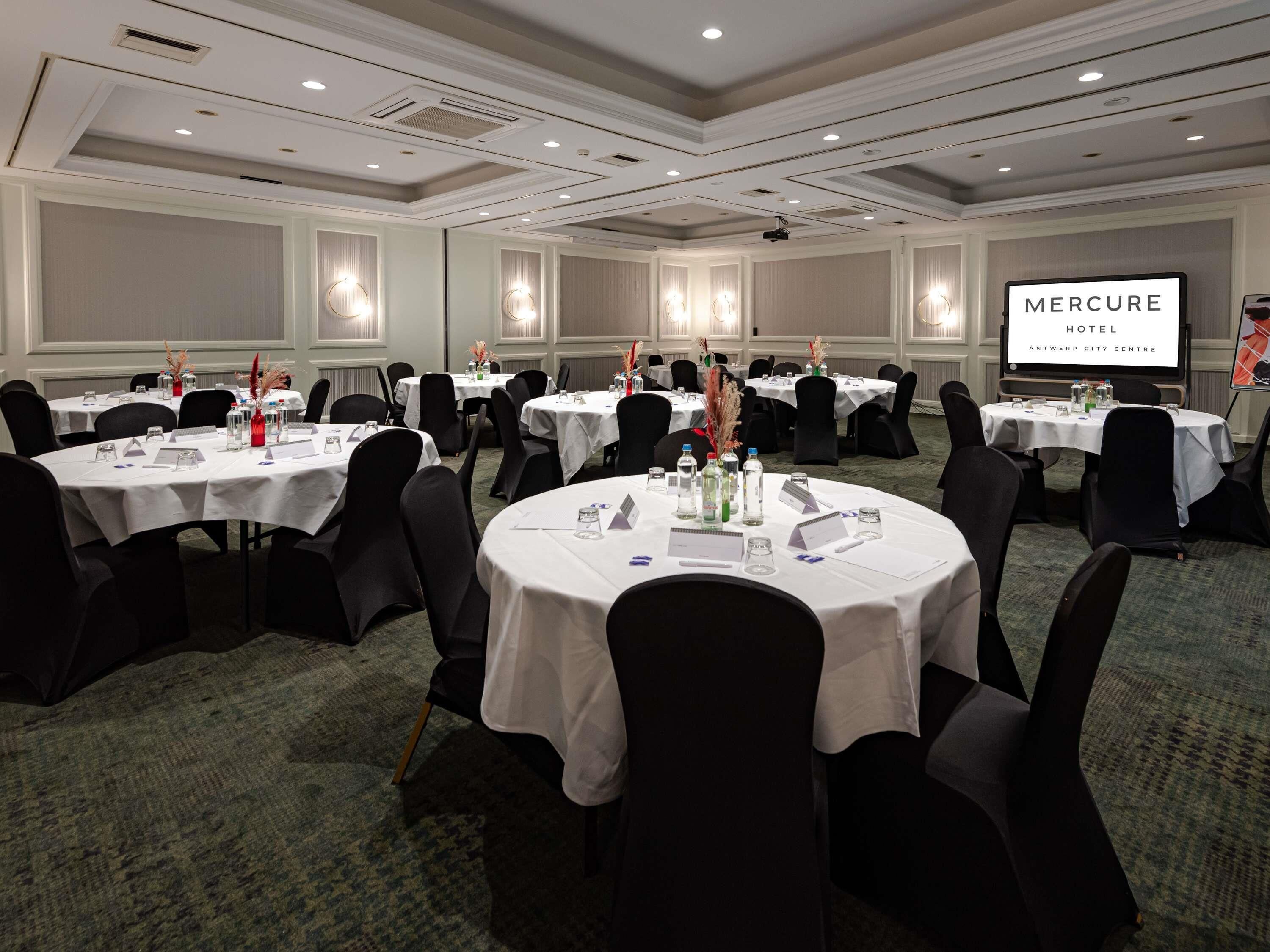 A conference room with round tables, black chairs, white tablecloths, and a projector screen displaying 'Mercure Hotel Antwerp City Centre'.