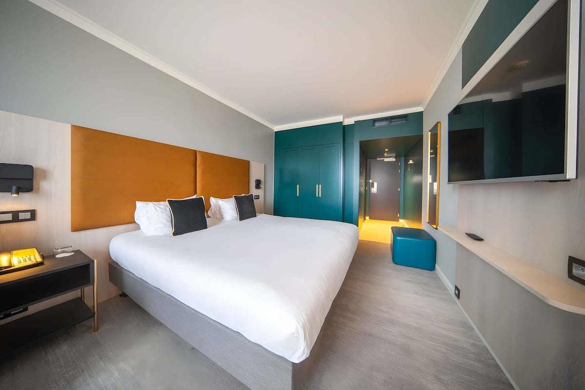 A modern hotel room with a large bed, a bedside table with a lamp, a TV mounted on the wall, and a wardrobe.