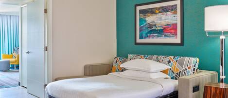 A hotel room with a bed, a teal accent wall, a framed abstract painting, and a bedside lamp.
