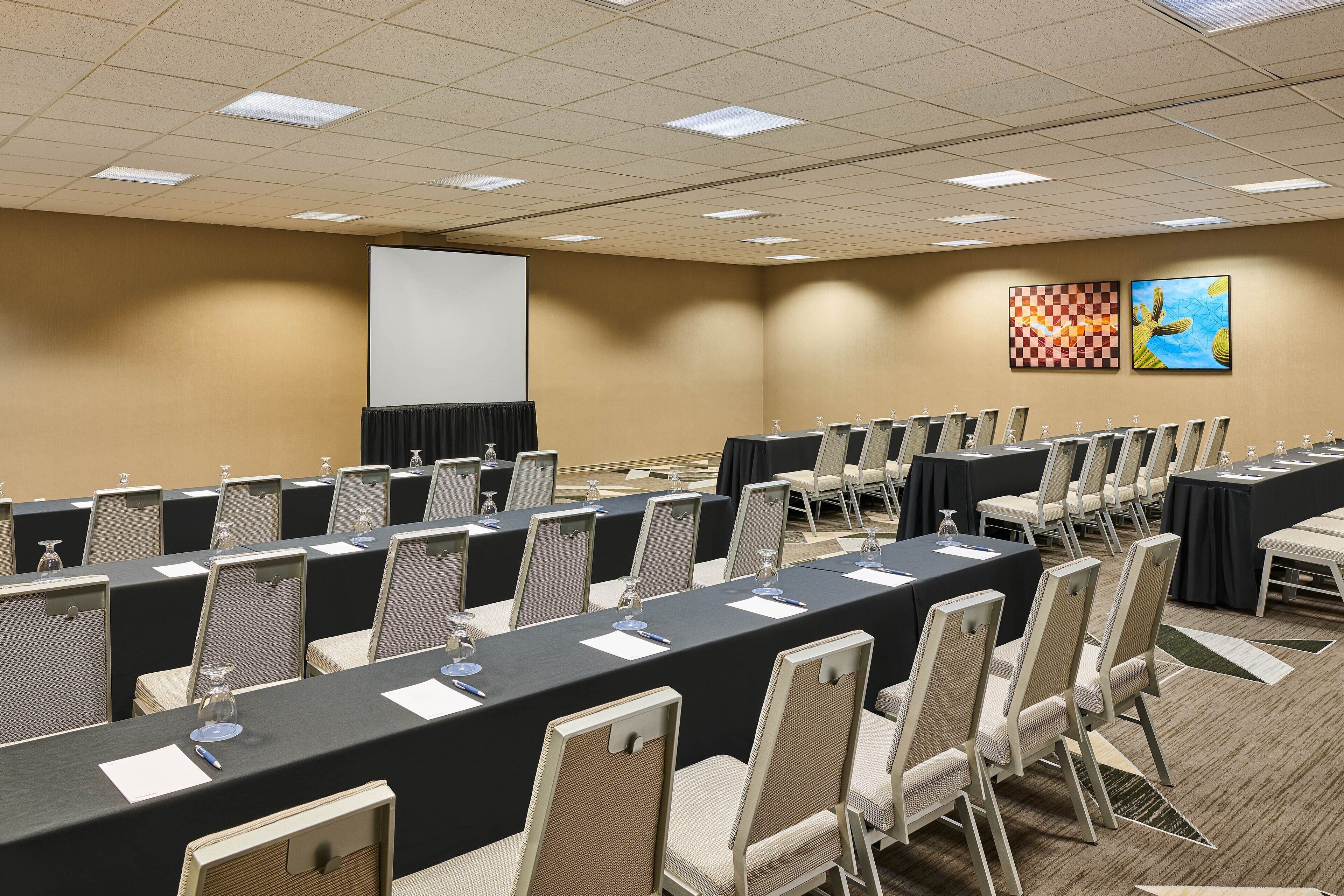 A conference room with long tables, chairs, a projector screen, and two wall-mounted artworks.
