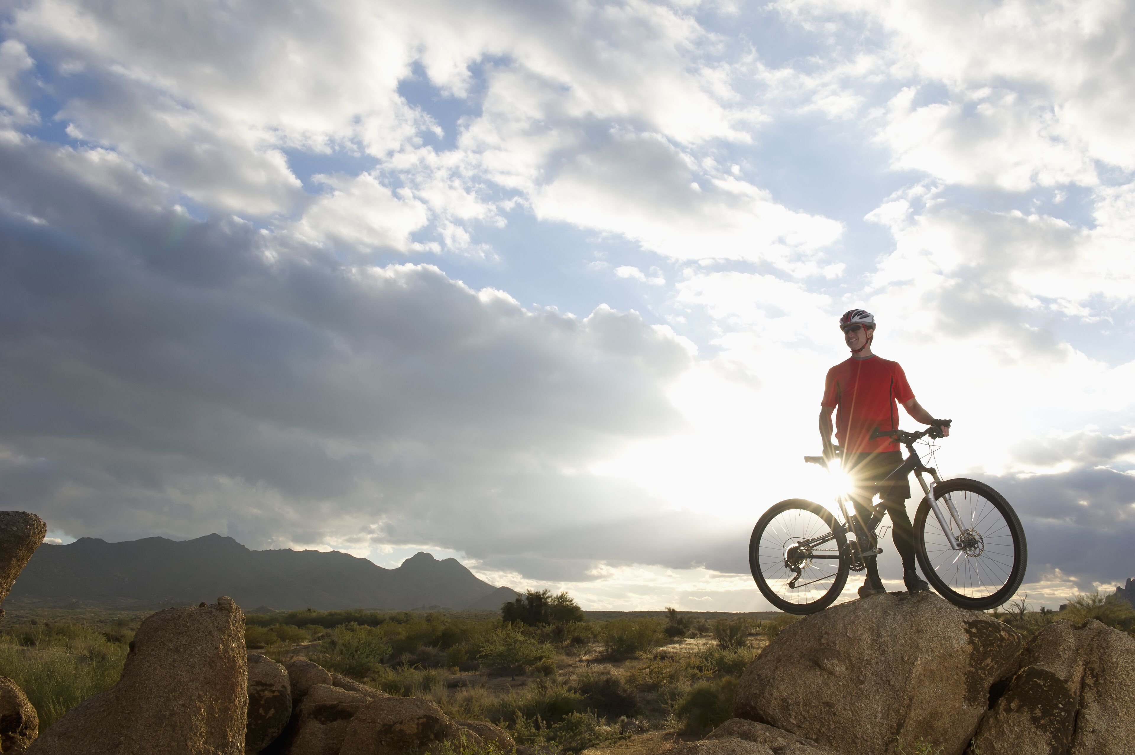 A person standing on a rock with a bicycle, with mountains and a cloudy sky in the background.