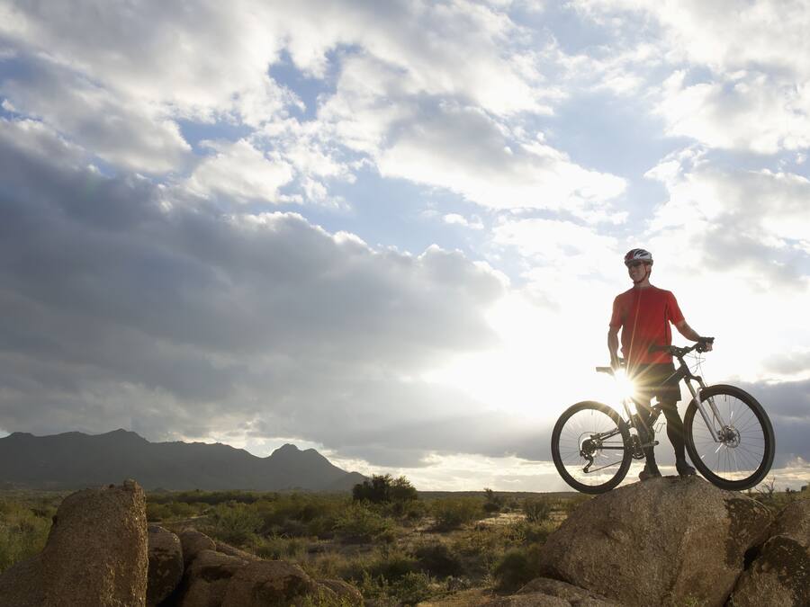 A person standing on a rock with a bicycle, with mountains and a cloudy sky in the background.