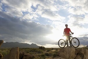 Una persona parada sobre una roca con una bicicleta, con montañas y un cielo nublado al fondo.