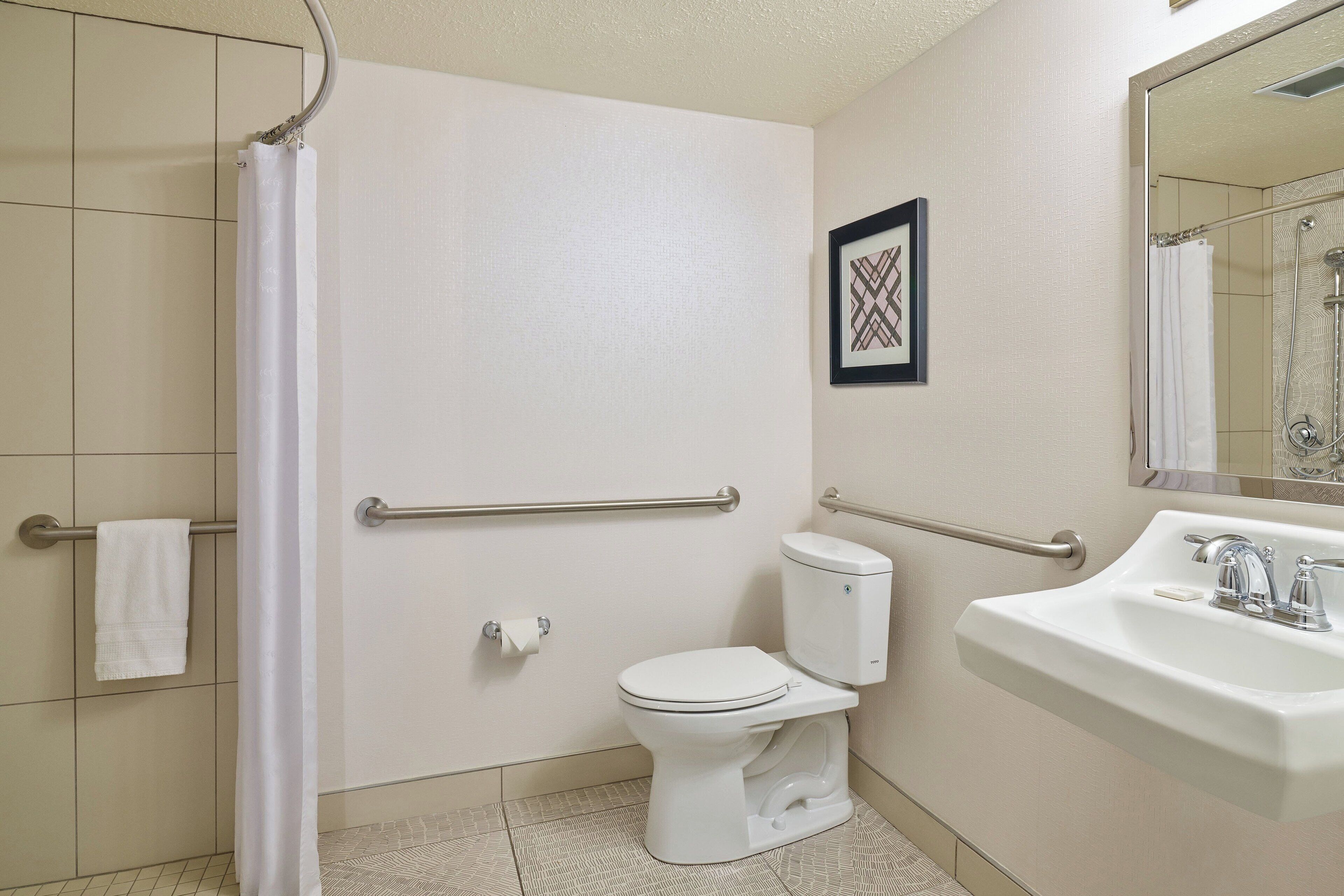Une salle de bain avec des toilettes blanches, un lavabo et un espace douche. Des barres d’appui ont été installées pour faciliter l’accès.