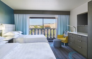 Habitación de hotel con dos camas, un escritorio, una silla, un televisor y un balcón con vistas.