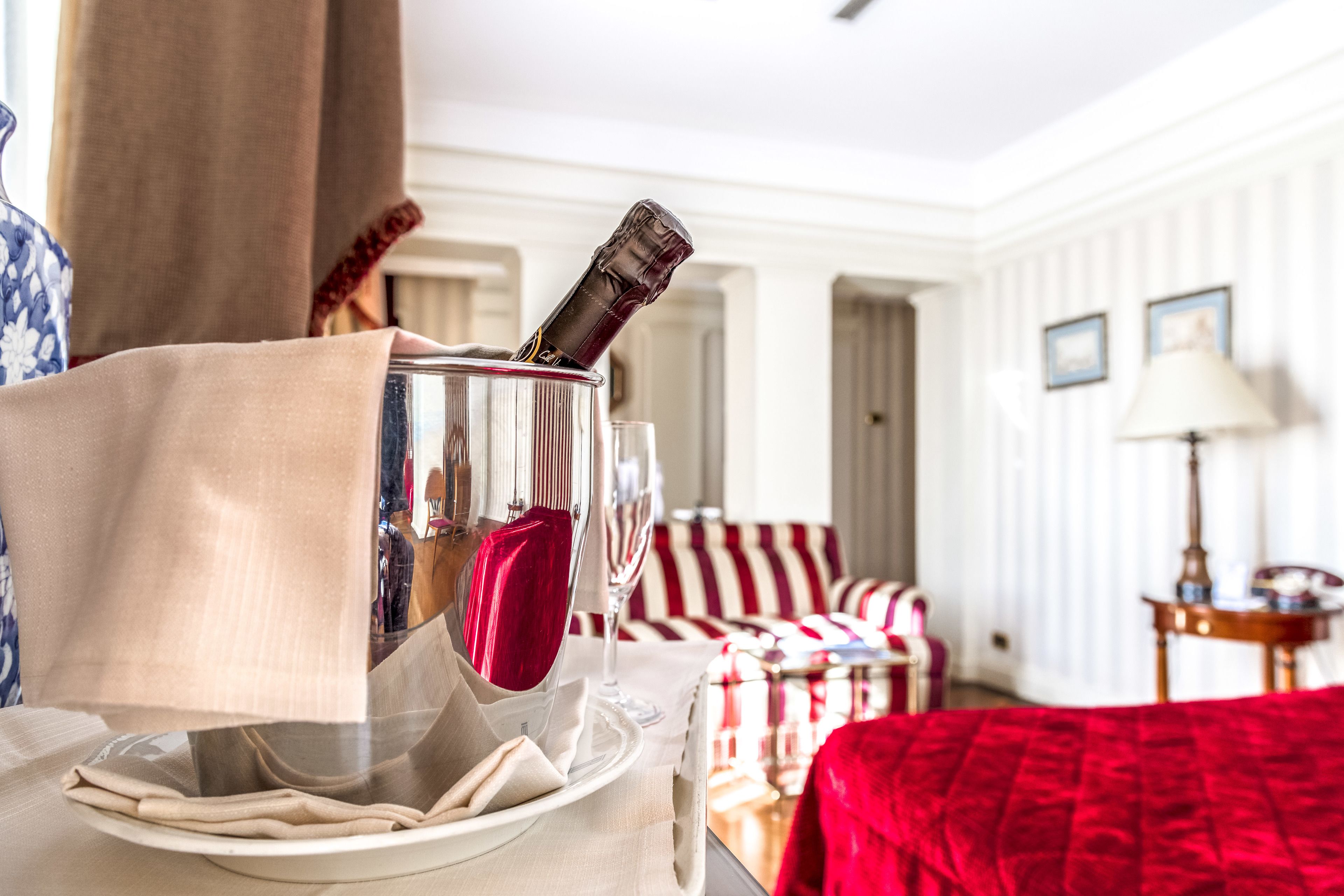 Habitación de hotel con una mesa de comedor dispuesta con una botella de vino, una copa y un plato. Se observa un sofá a rayas y una manta roja al fondo.