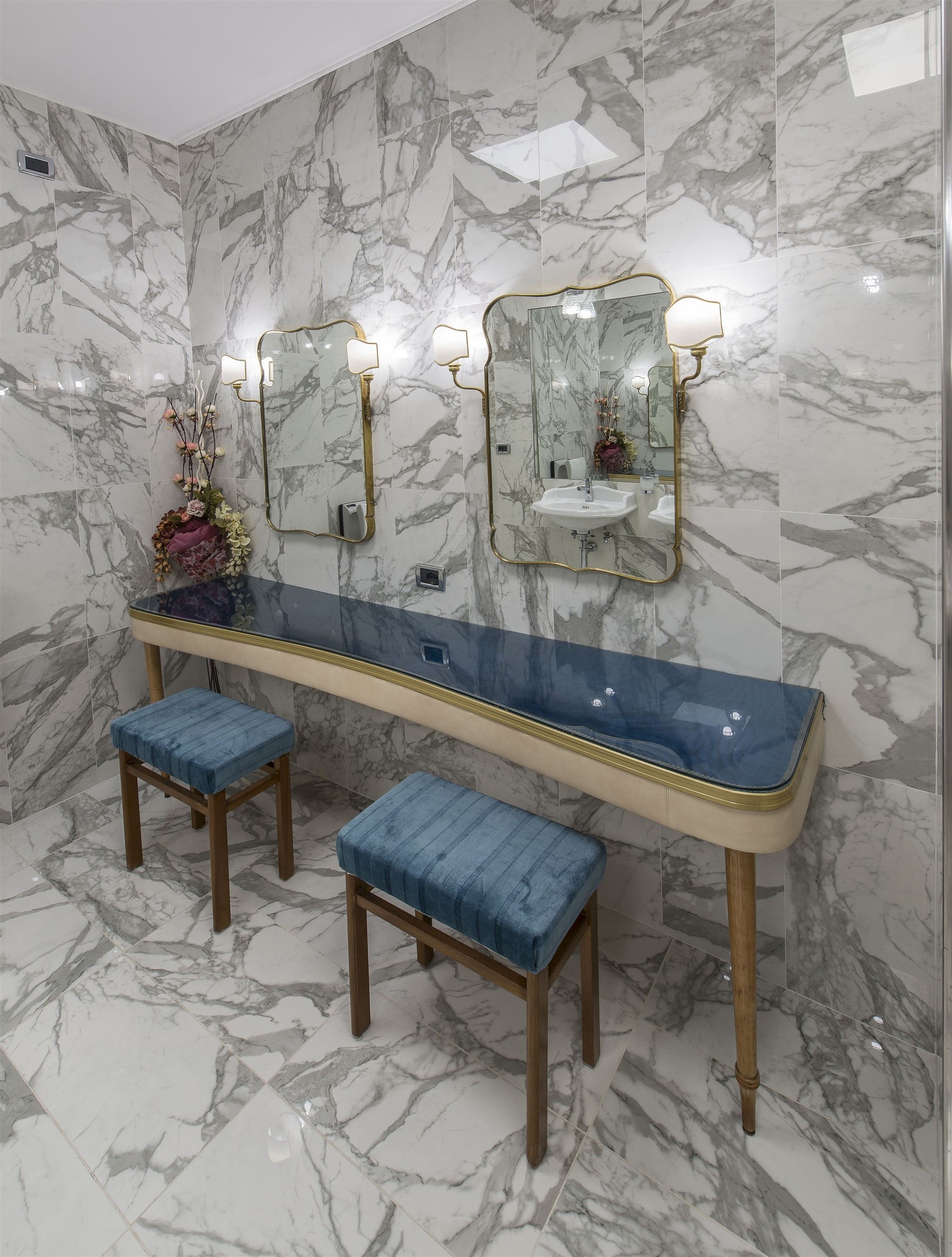 Un baño con paredes de mármol, un tocador largo con dos espejos y dos taburetes de madera.