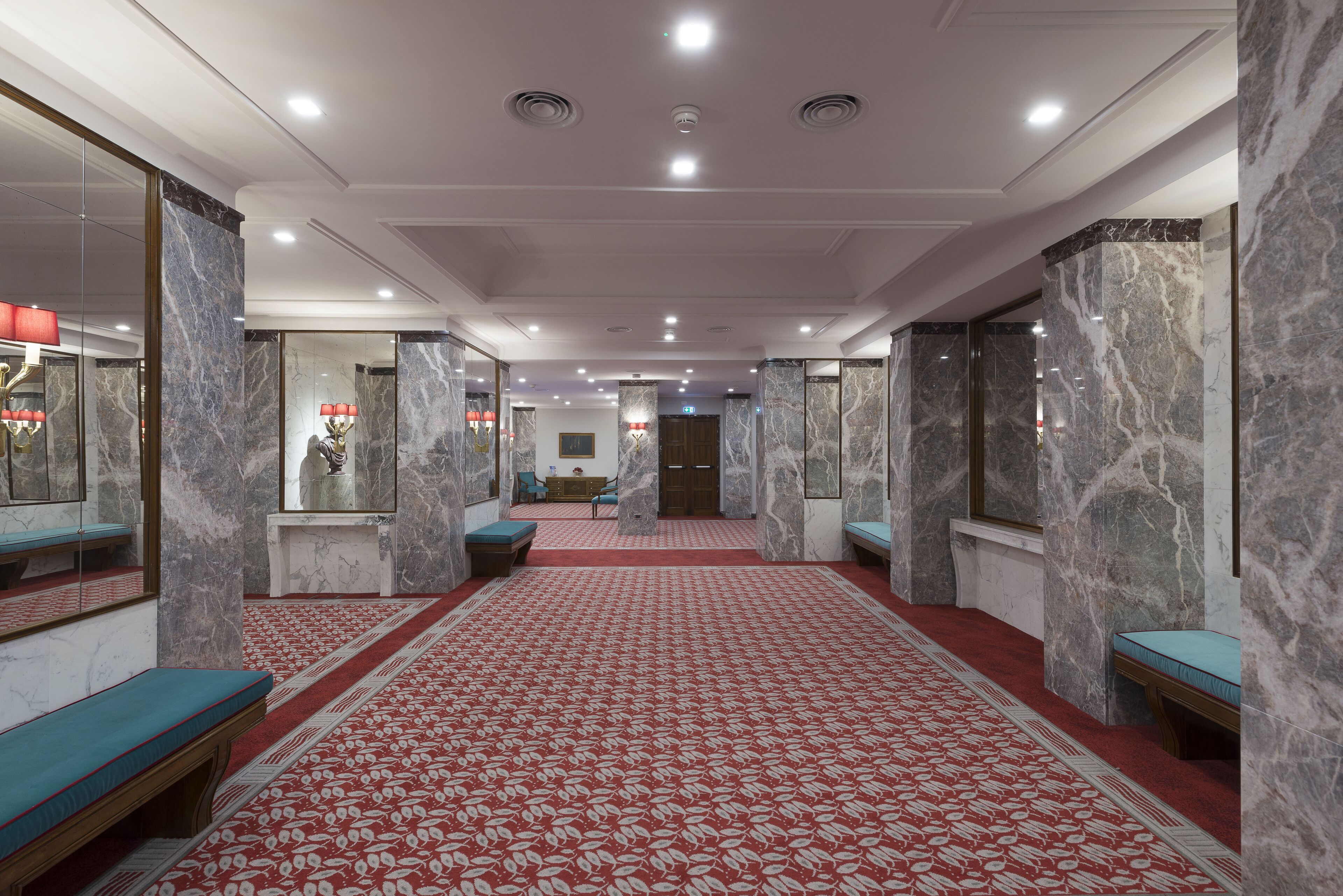 A hotel lobby with marble walls, red patterned carpet, and seating areas.