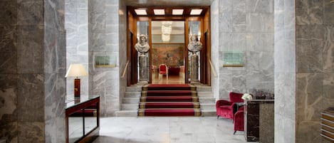Un vestíbulo con suelo de mármol y una escalera con alfombra roja, una recepción y una silla roja.