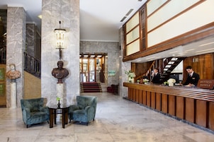 A hotel lobby with marble columns, a reception desk, and a staircase leading to an upper floor.