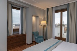 A hotel room with a large bed, a chair, a lamp, and a view of the city through the windows.