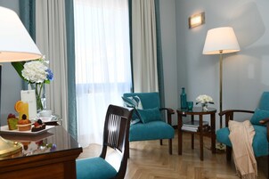 Habitación de hotel con piso de madera, sillones turquesa, una mesita de madera, una mesa de centro de vidrio, una lámpara y un florero con flores.