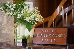 A wooden sign with "HOTEL MEDITERRANEO ROMA" text, a vase with white flowers, and a small potted plant on a marble surface.