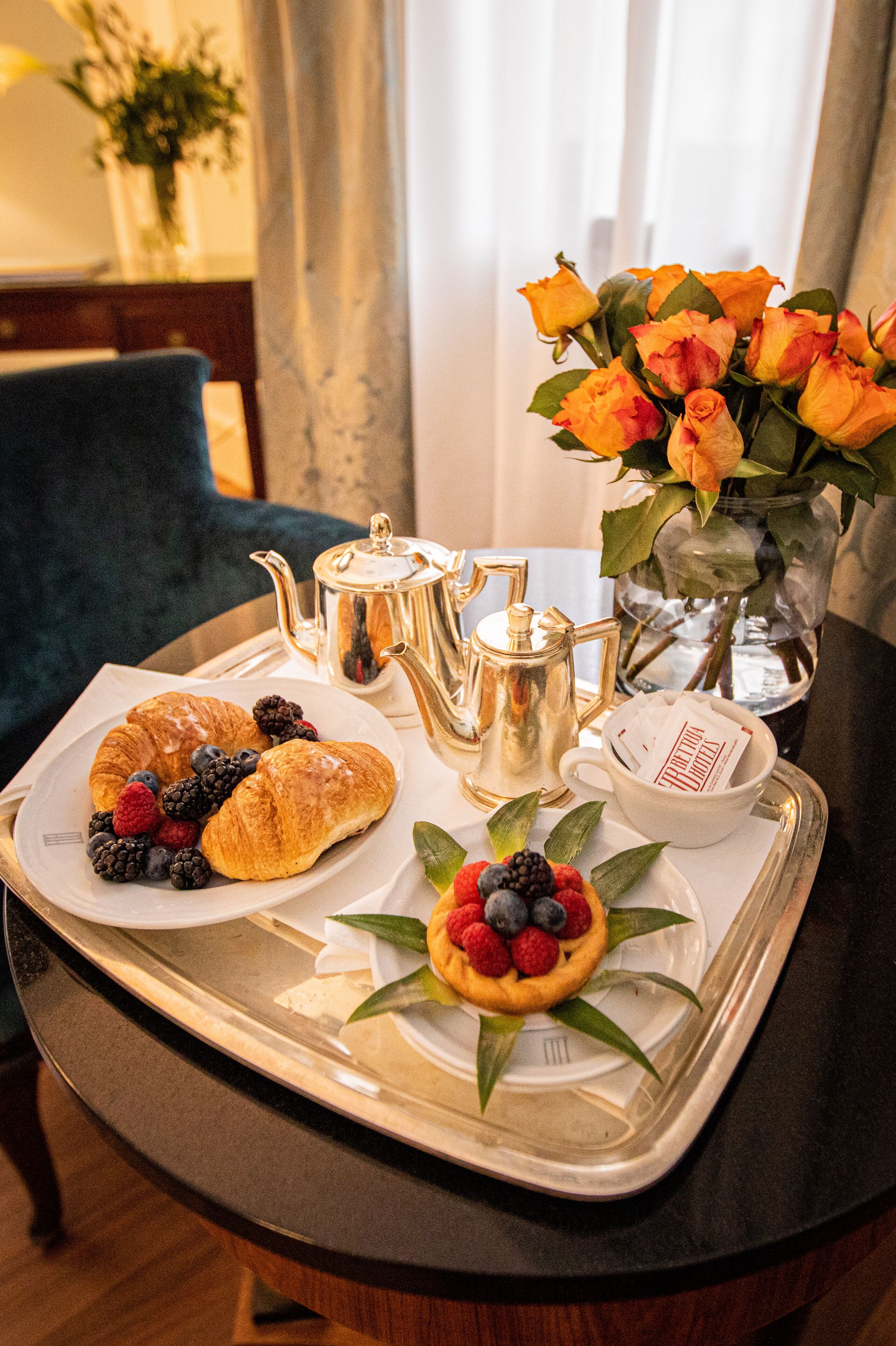 A tray with croissants, berries, a small fruit tart, a teapot, a cup, and a vase of flowers.
