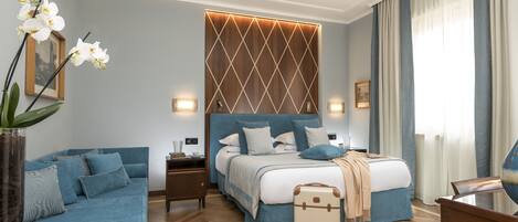 Una habitación de hotel moderna con una cama grande, un sofá azul, una mesita de noche de madera, una mesilla auxiliar y una mesa de centro de vidrio.