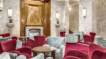 A luxurious lobby with marble walls, red and grey upholstered chairs, a wooden fireplace, and wall-mounted sconces.