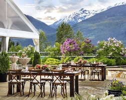 Un área de comedor al aire libre con mesas y sillas, rodeada de plantas y flores en macetas, y una vista de la montaña al fondo.