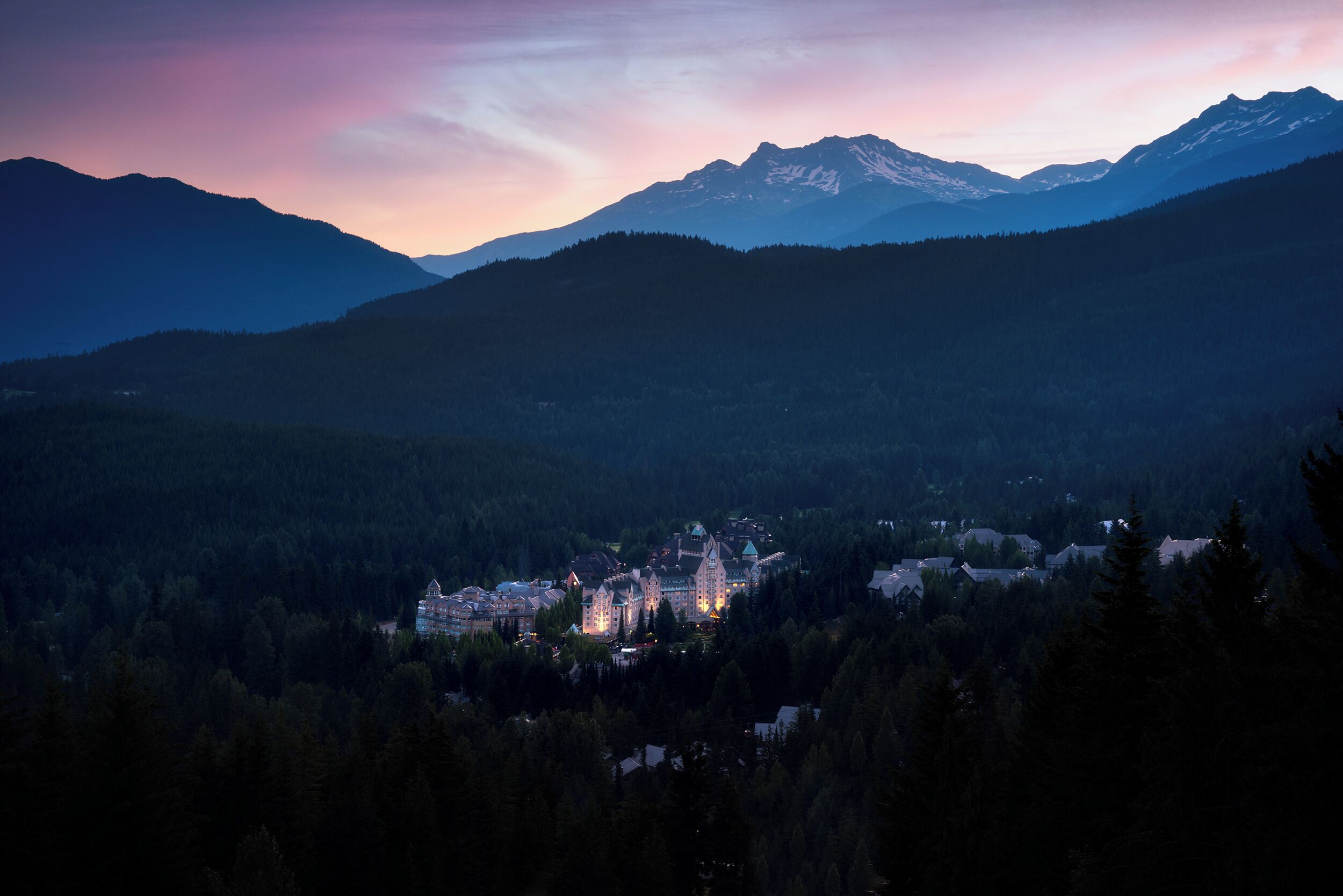 A town with a prominent building featuring a tower, surrounded by forested hills and mountains.