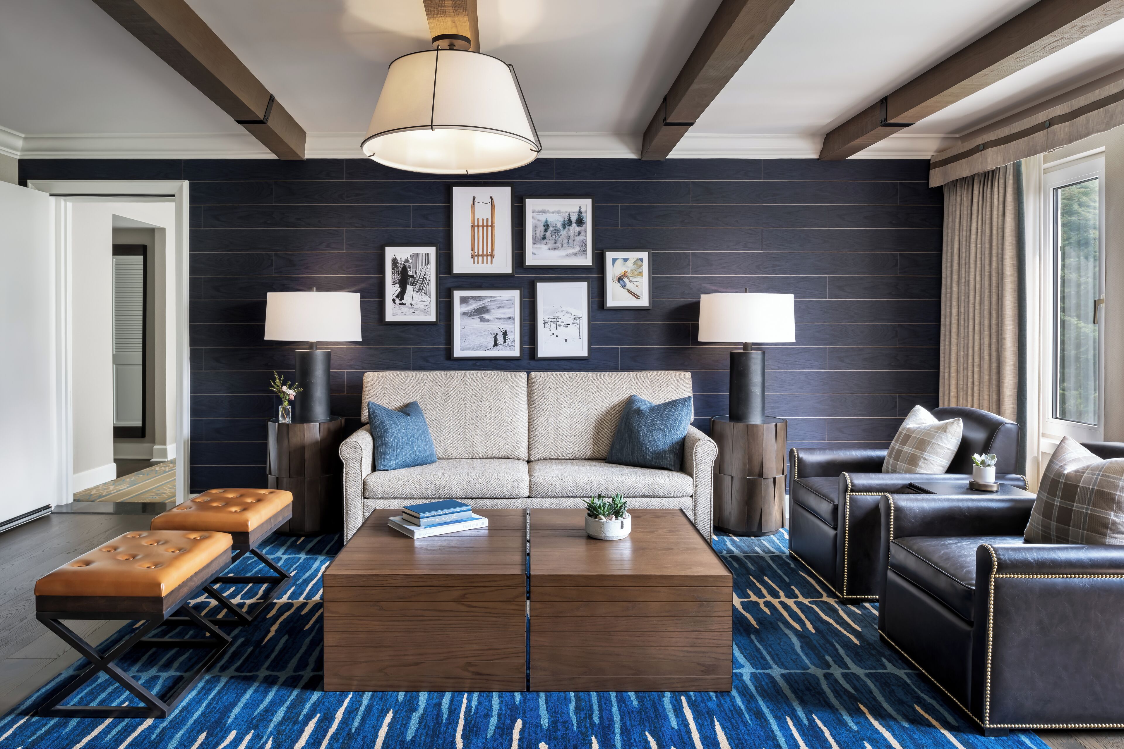 Ein modernes Wohnzimmer mit dunkler Wand, einem beige farbenen Sofa, zwei schwarzen Ledersofas, einem Holztisch und einem blau gemusterten Teppich.