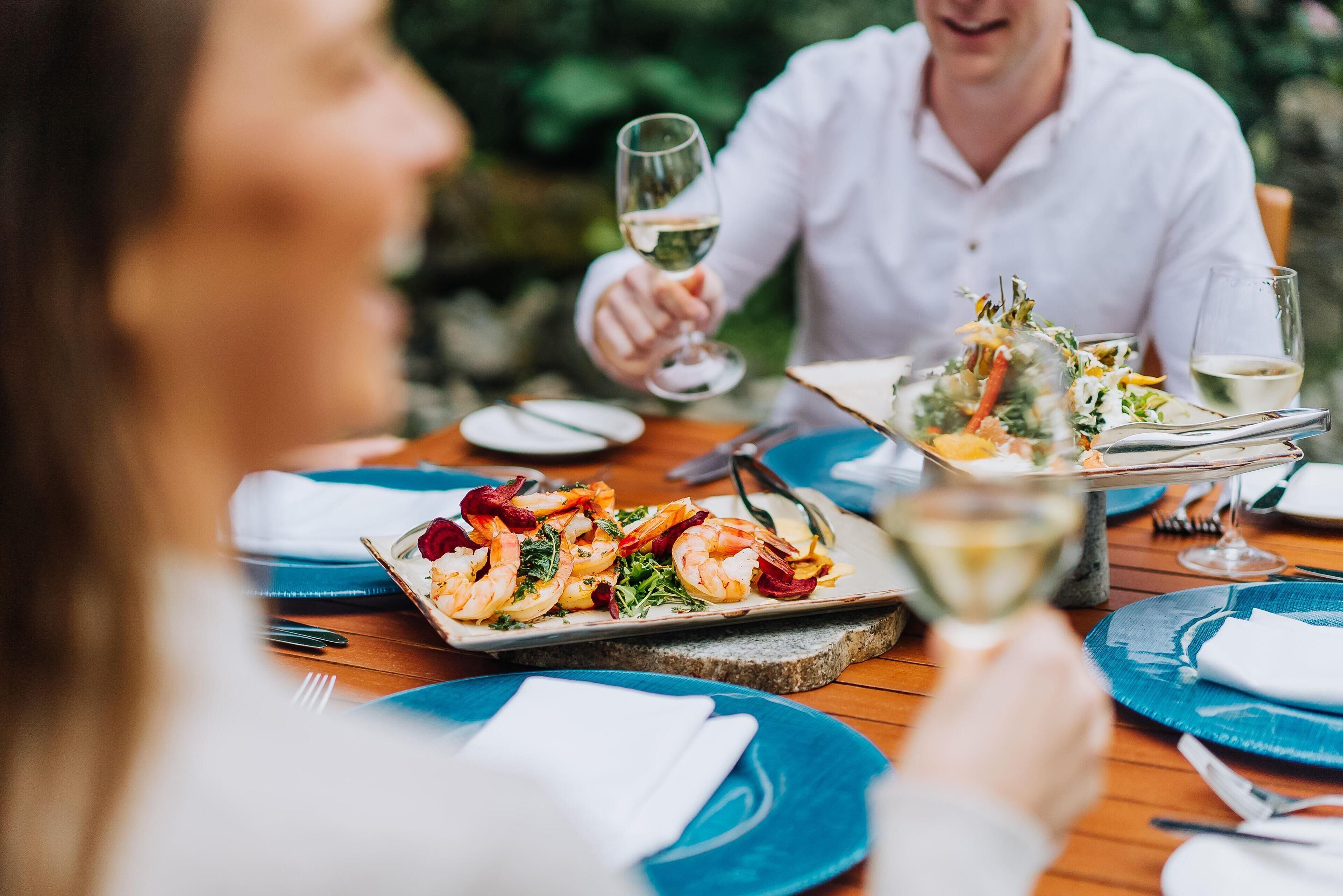 Una mesa de comedor con platos de comida, una copa de vino y una persona sosteniendo un vaso.
