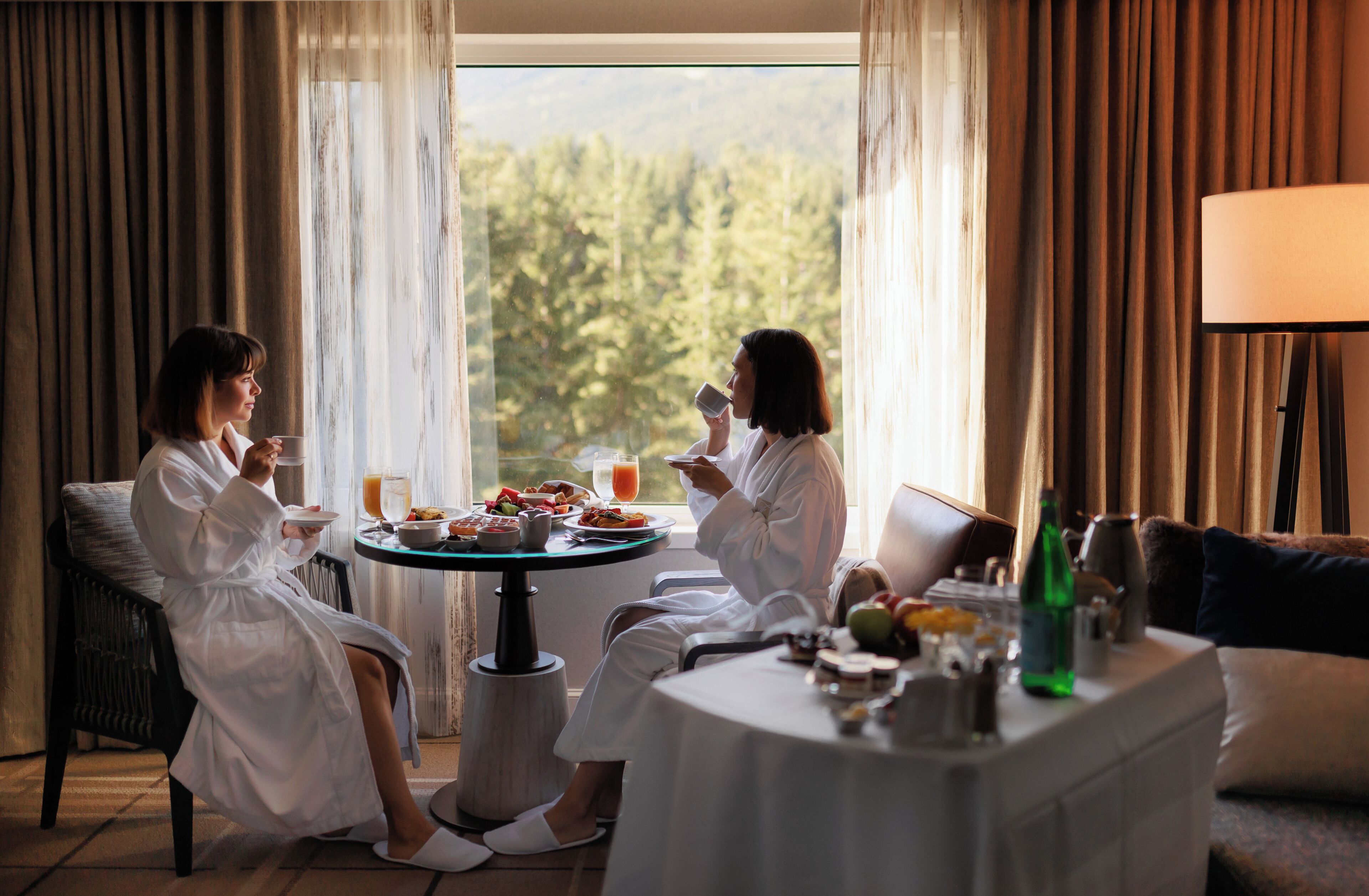 A room with two people seated at a table with breakfast, a lamp, and a view of trees through the window.