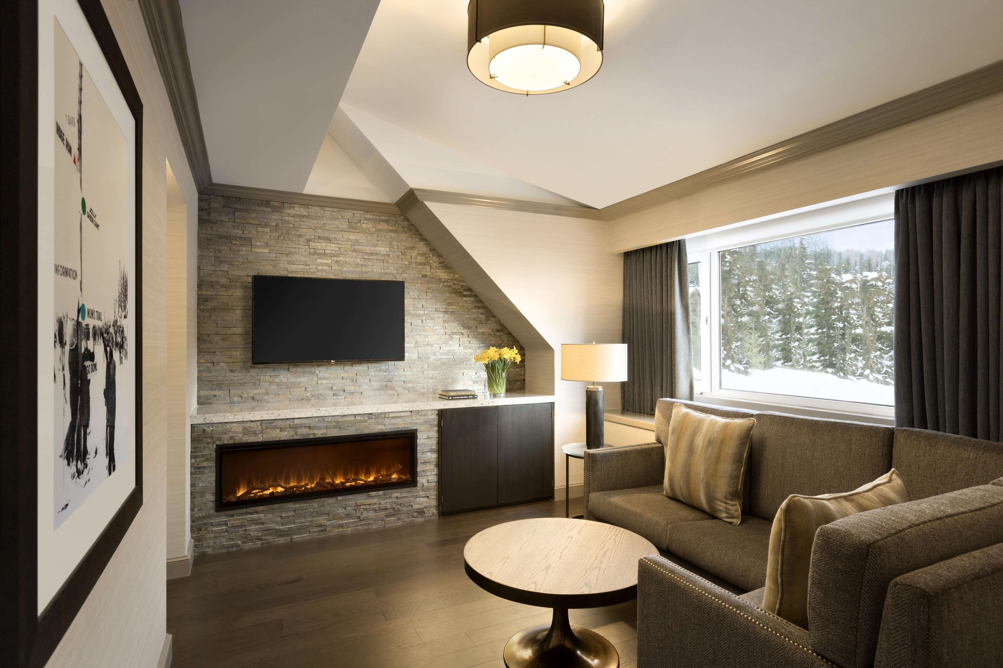 A modern living room with a fireplace, a flat-screen TV mounted on a stone wall, a sofa, a round coffee table, and a large window with a view of snow-covered trees.