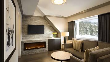 A modern living room with a fireplace, a flat-screen TV mounted on a stone wall, a sofa, a round coffee table, and a large window with a view of snow-covered trees.