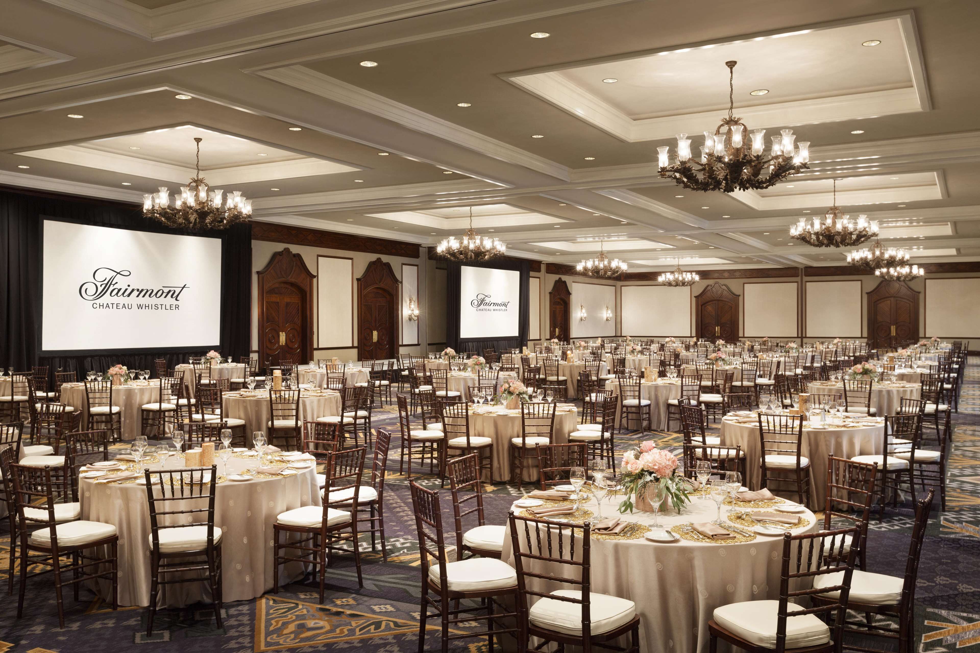 A banquet hall with round tables, chairs, chandeliers, and a large screen displaying the hotel's name.