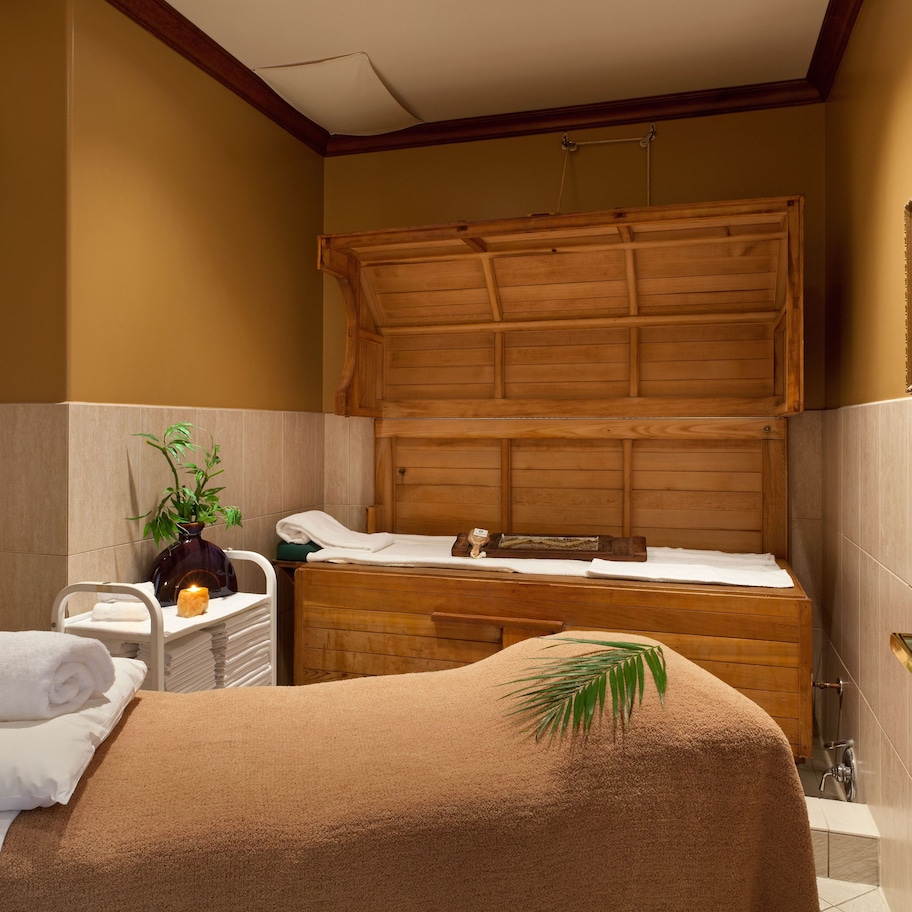 A spa room with a wooden bench, a small table with a plant, and a towel on the bed.