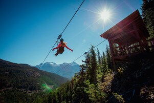 A person zip-lining over a forested mountain area with a wooden structure on the right.