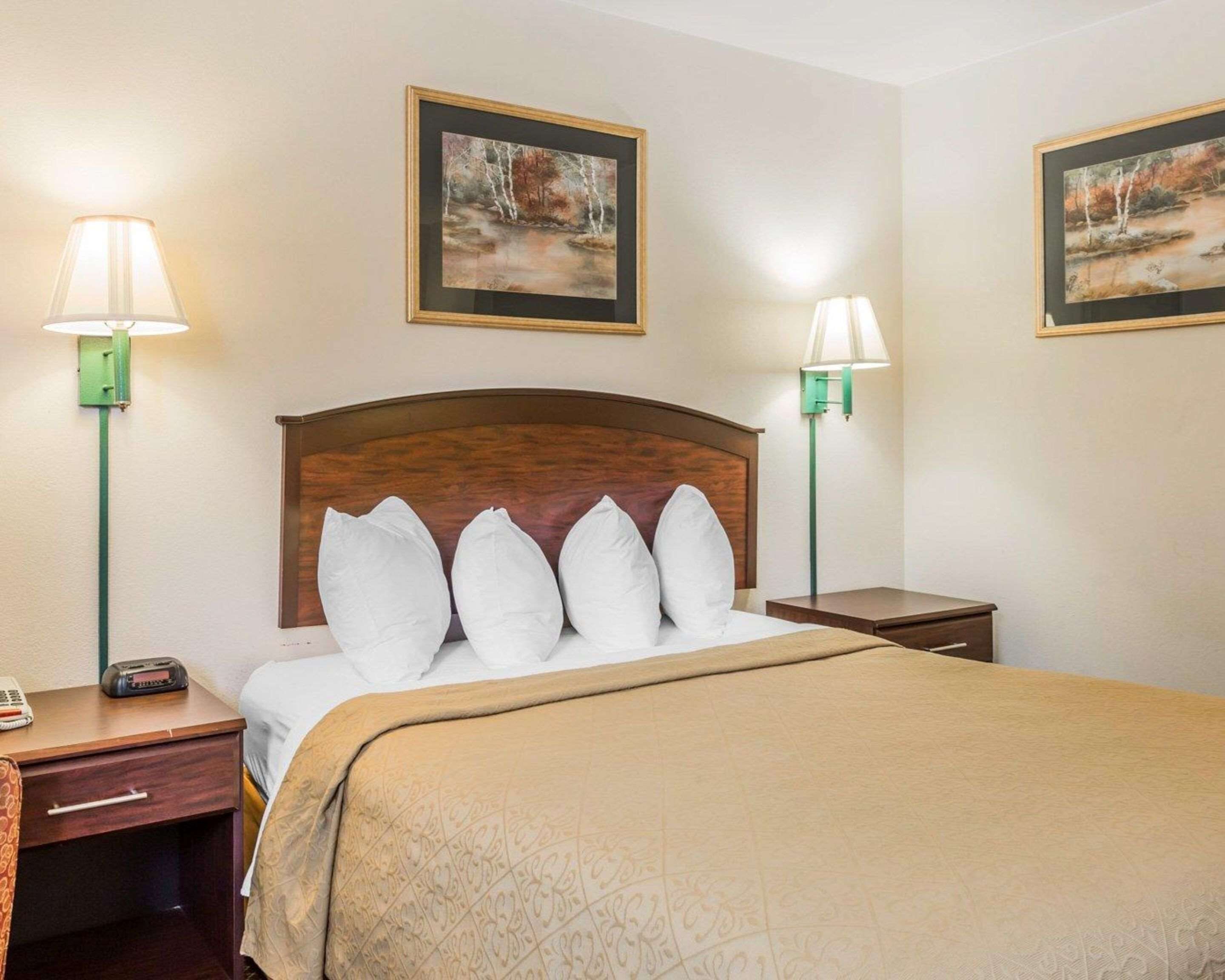Une chambre d’hôtel avec une tête de lit en bois, un lit avec des oreillers blancs, deux tables de chevet et deux lampes fixées au mur.