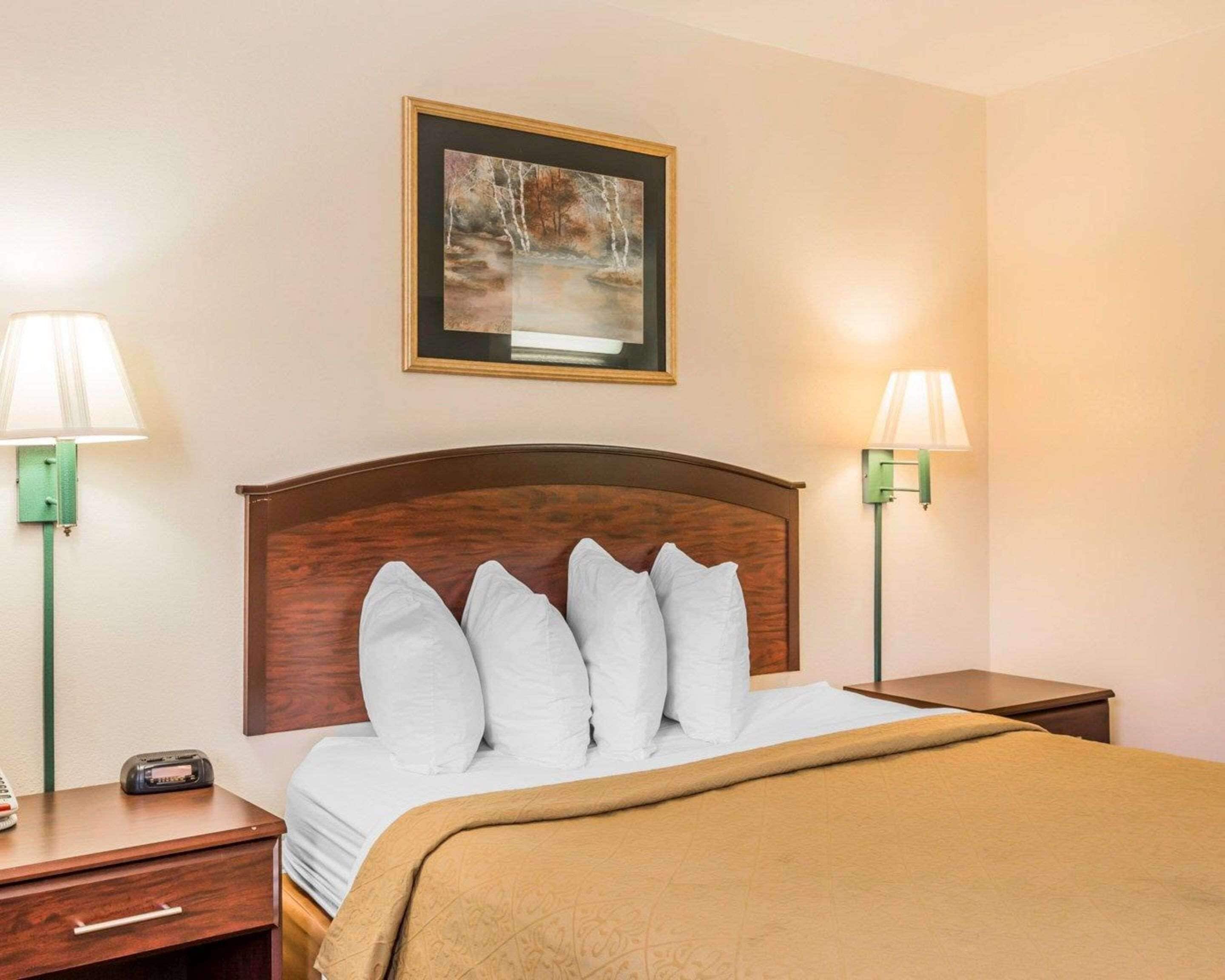 A hotel room with a wooden headboard, white bedding, a framed painting above the bed, and two bedside lamps.