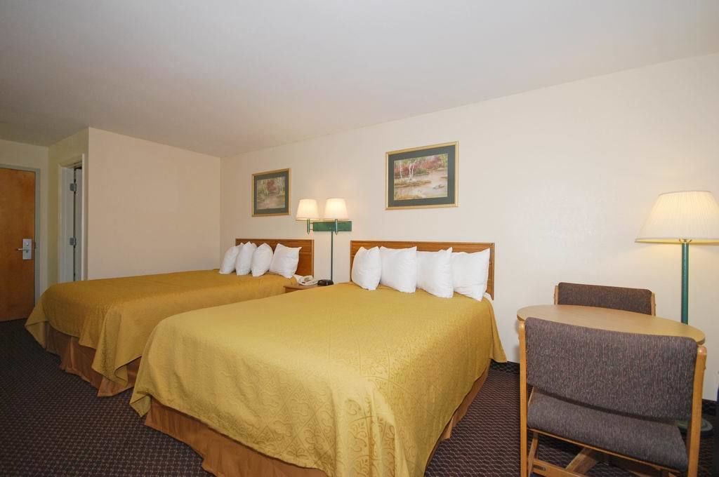 A hotel room with two beds, a chair, a table, and two wall-mounted lamps.