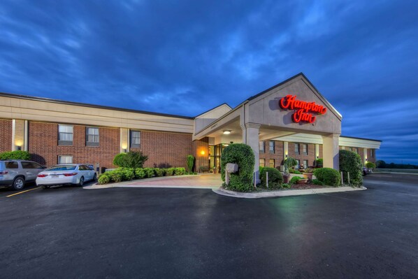 A Hampton Inn with a brick facade, a well-lit entrance, and a parking lot with several cars.