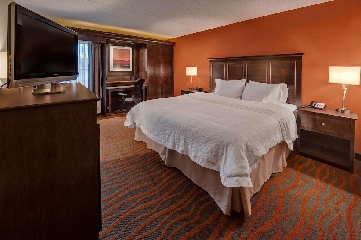 A hotel room with a large bed, a desk with a TV, two bedside lamps, and a patterned carpet.
