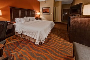 A hotel room with a large bed, a desk with a lamp, a television, and a patterned carpet.