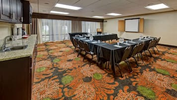 A conference room with a carpeted floor, a kitchen area with a sink, and a projector screen on the wall.