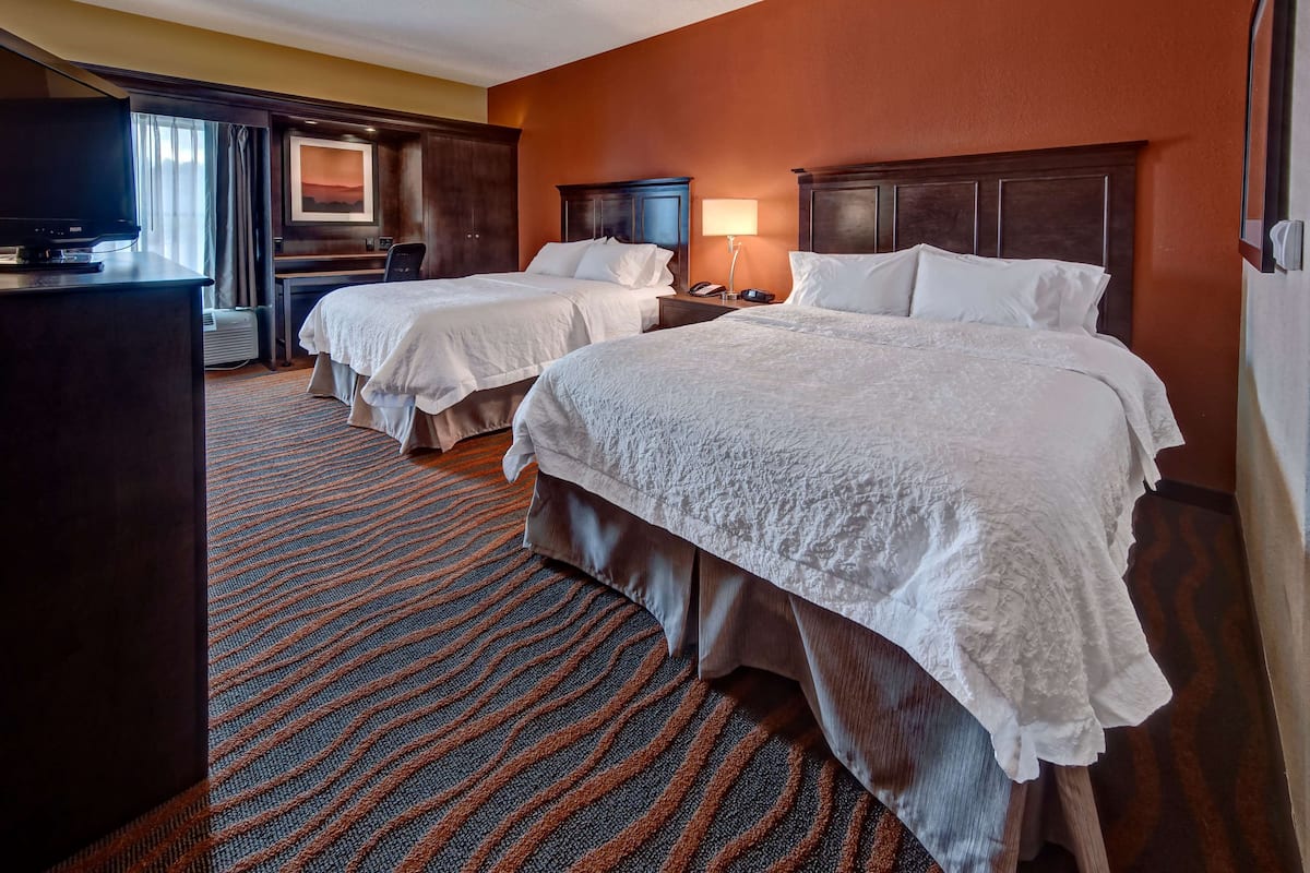 A hotel room with two beds, a desk, a TV, and a large window with curtains.