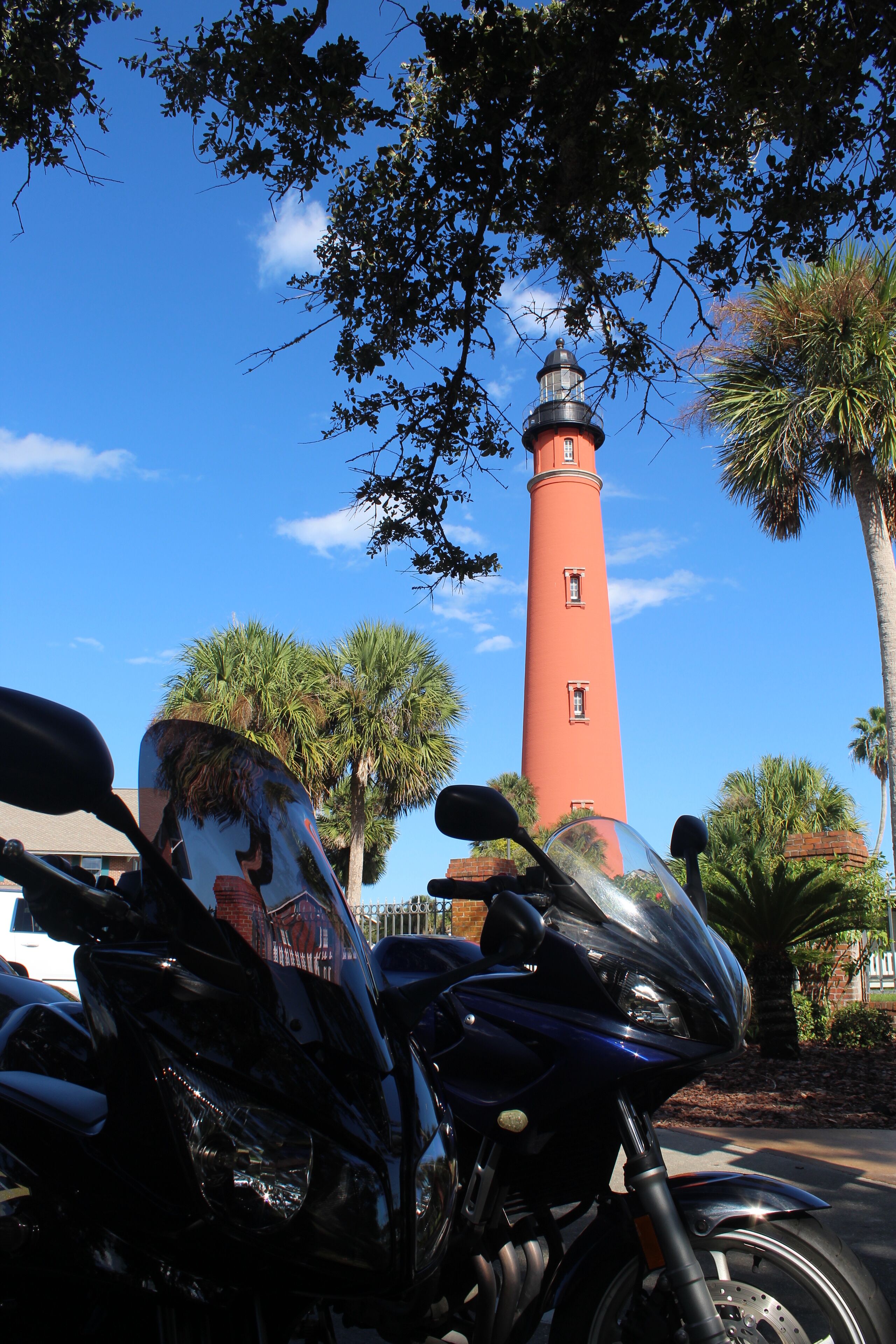 A motorcycle parked in front of a red lighthouse with palm trees and a clear blue sky in the background.